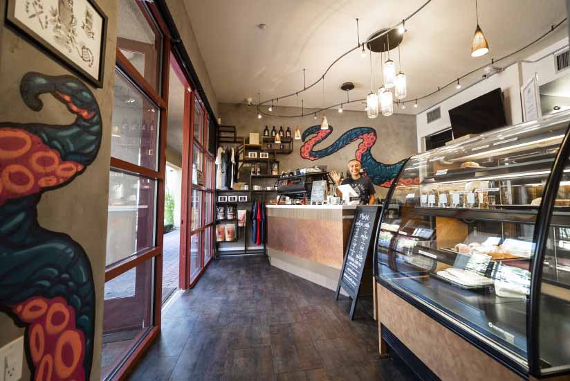 Inside of Seven Seas Roasting Aliso Viejo Location with one barista at the counter waving at the camera. 