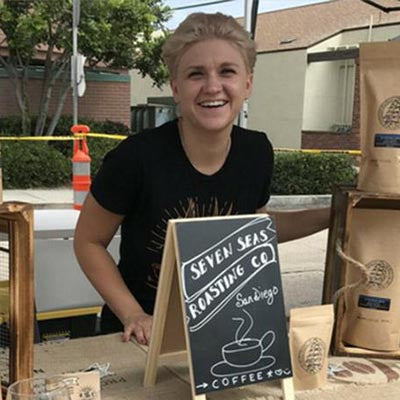 Sasha D, caucasian female wearing blackshirt smiling behind farmers market table with seven seas roasting sign ontop of table  